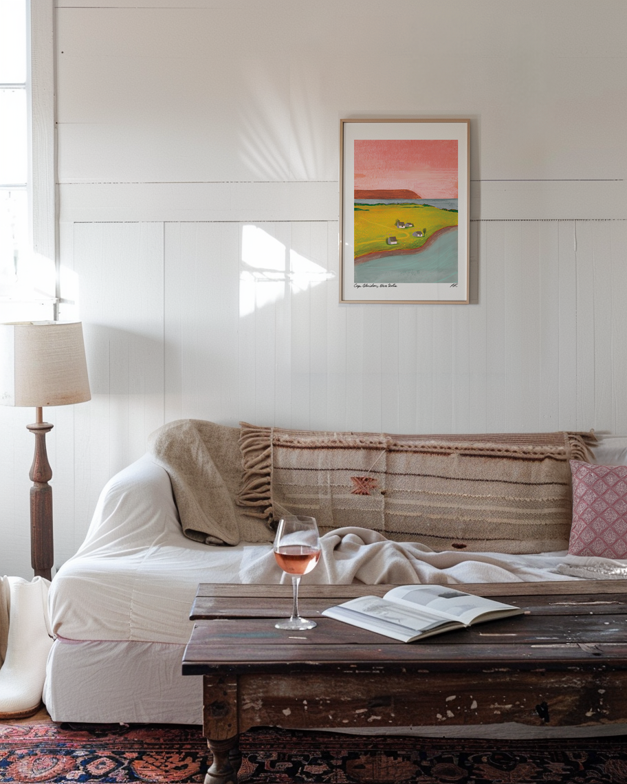 A bohemian white living room with white painted wooden walls features the Wild Rock Rosé print in a light wooden frame. The couch looks inviting with natural textures of blankets and a cotton cover. A glass of rosé sits on the shabby chic coffee table.