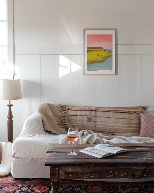 A bohemian white living room with white painted wooden walls features the Wild Rock Rosé print in a light wooden frame. The couch looks inviting with natural textures of blankets and a cotton cover. A glass of rosé sits on the shabby chic coffee table.