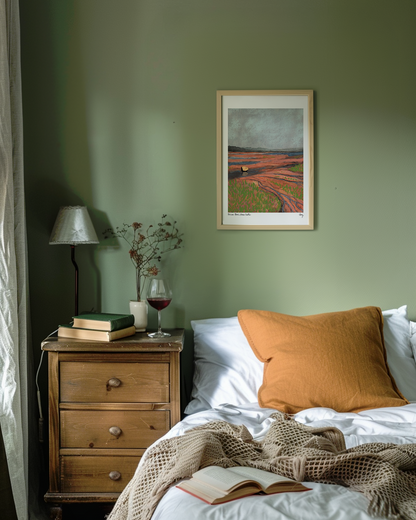 A cozy bedroom with a sage green accent wall features the Wild Rock Red art print framed behind the bed. A glass of red wine sits on the boho nightstand along with books and a lamp. Light comes in from the window.